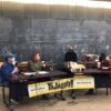 the YaJagoff podcast crew seated at tables with their black and gold logo banner, recording the episode in the community room at Construction Junction in from of the blackboard wall.