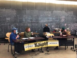 the YaJagoff podcast crew seated at tables with their black and gold logo banner, recording the episode in the community room at Construction Junction in from of the blackboard wall.
